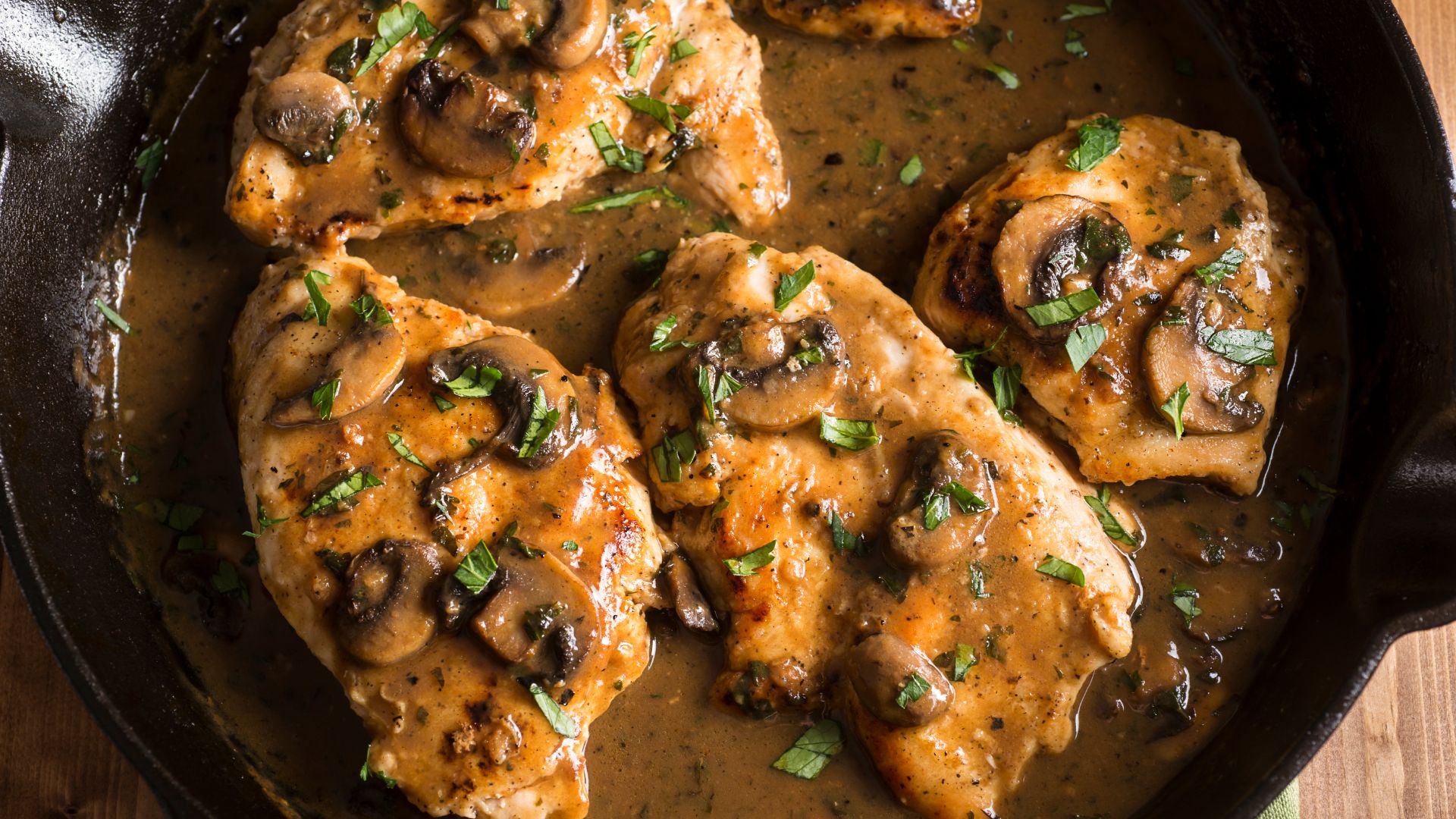 Top-down view of a classic Chicken Marsala in a cast iron skillet, with golden-brown chicken pieces simmered in a rich, dark gravy, garnished with mushrooms and fresh parsley.