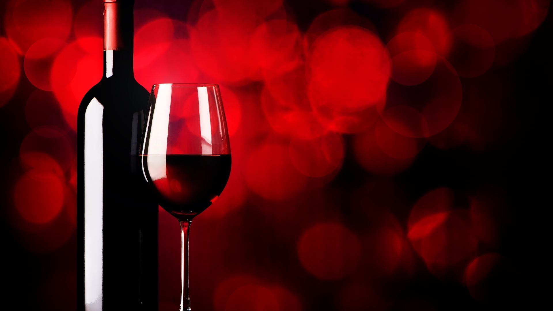 A bottle of red fortified wine stands next to a glass filled with deep ruby wine, set against a vibrant red bokeh background.