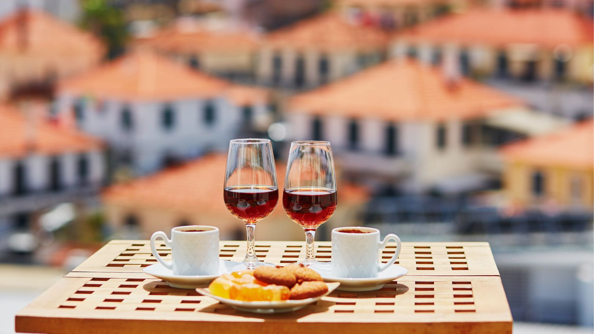 Two glasses of amber-hued Madeira wine on a wooden table, accompanied by two cups of espresso and a plate of biscuits and candied fruit, with a backdrop of orange-tiled rooftops in Madeira, Portugal.