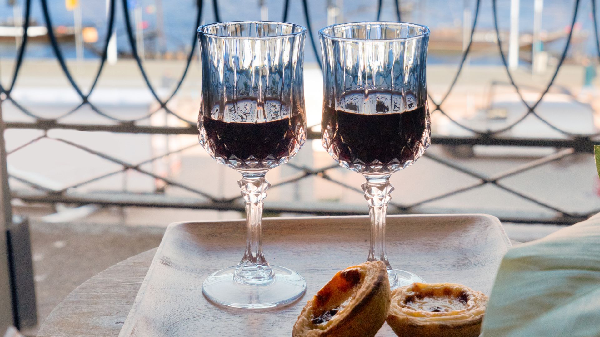 Two crystal glasses filled with deep red port wine on a wooden tray, accompanied by Portuguese custard tarts, set against a scenic waterfront view.