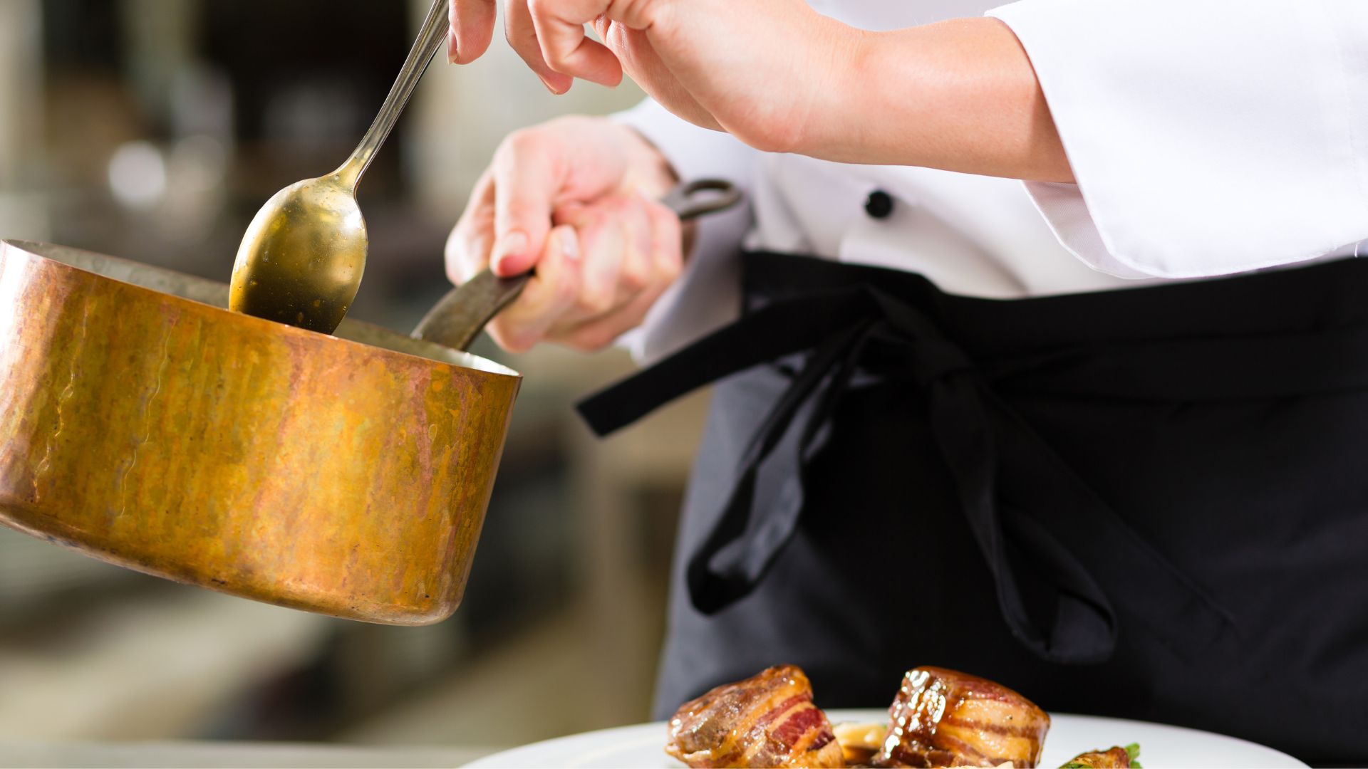 A saucier in a white chef's coat and black apron pours sauce from a copper saucepan onto plated food.