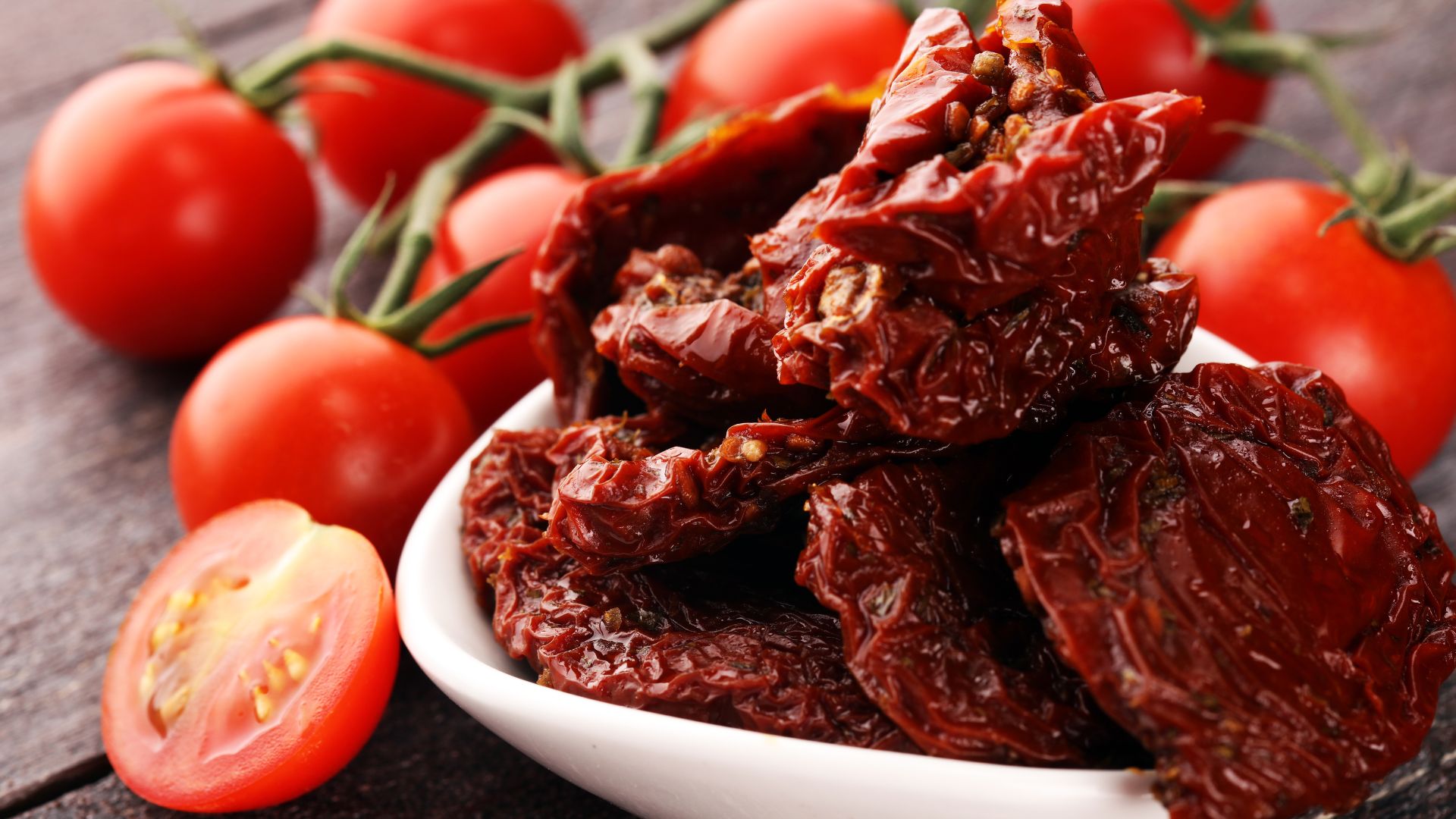 A bowl of sun-dried tomatoes surrounded by fresh red tomatoes on the vine, with one sliced tomato in the foreground.