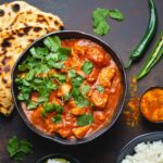 Bowl of chicken tikka masala in rich tomato sauce, garnished with cilantro, served with naan and rice.