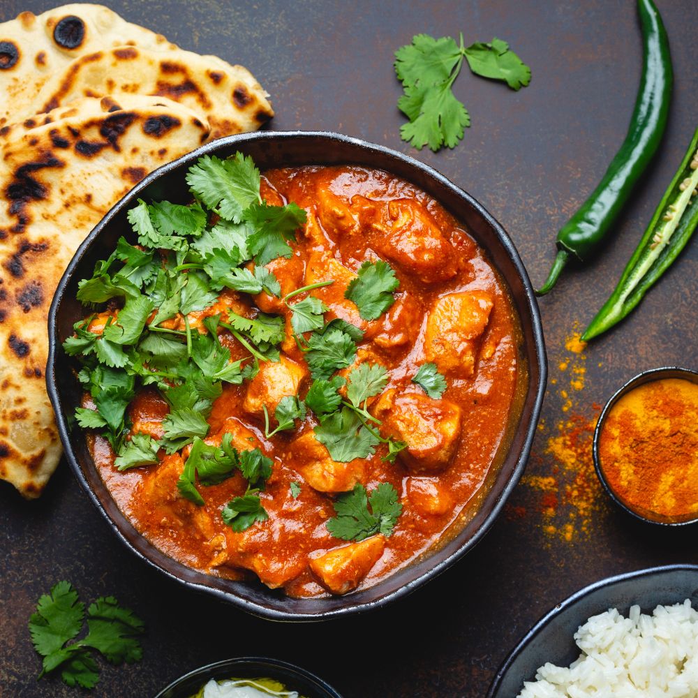 Bowl of chicken tikka masala in rich tomato sauce, garnished with cilantro, served with naan and rice.